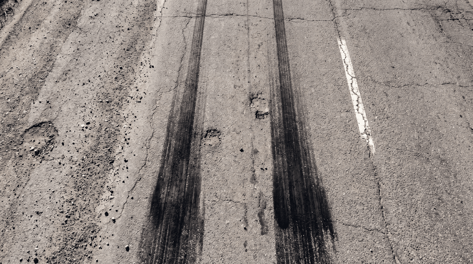 Senkrechte Aufnahme von trockenem Asphalt mit zwei parallelen schwarzen Bremsspuren, die zum unteren Bildrand hin dunkler und breiter werden (KI-generiertes Bild)